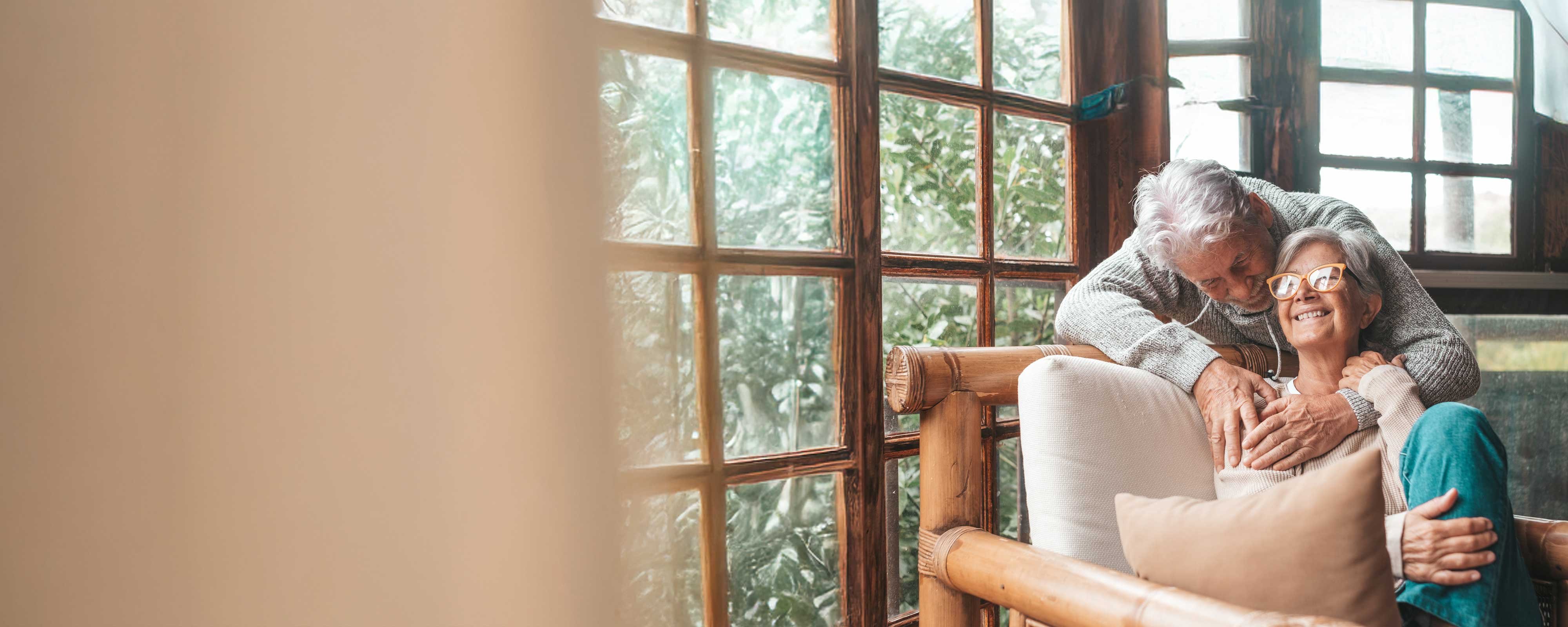 A senior couple sharing an embrace while one sits in a chair and the gentleman leans over the chair from behind.